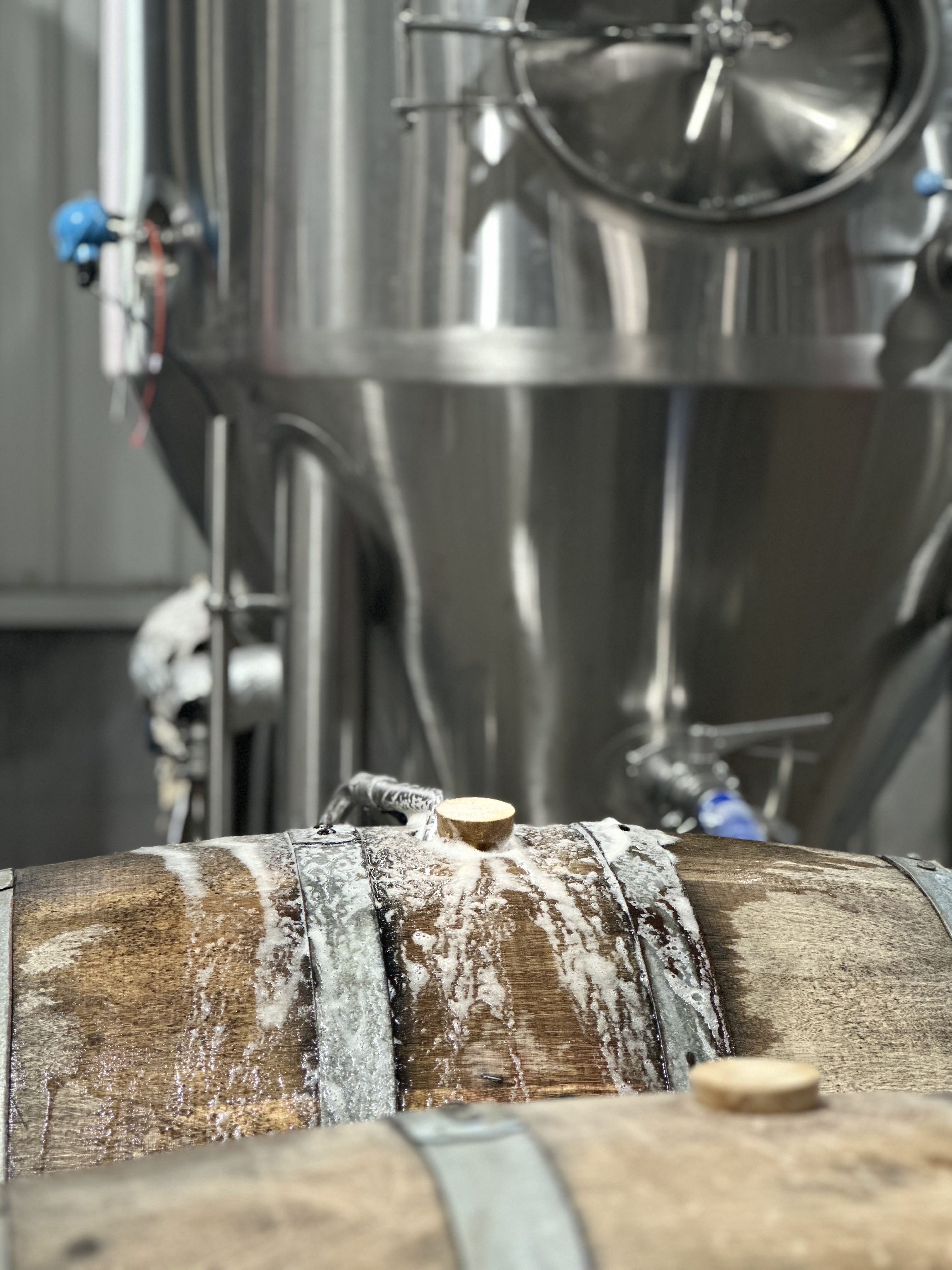 An oak barrel foaming over at the bung hole during fermentation, stainless tanks behind it